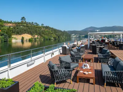 Sonniges Oberdeck der Douro Queen. Gäste entspannen in bequemen Sesseln. Eine grüne, hügelige Flusslandschaft zieht im Hintergrund vorbei. Tische mit Wein und Snacks sind zu sehen.