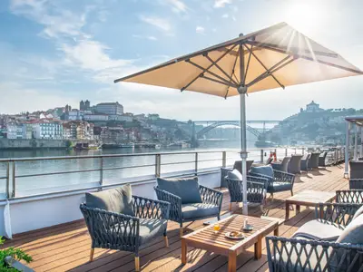 Panoramablick vom Sonnendeck eines Flusskreuzfahrtschiffs auf die bunte Skyline von Porto, den Fluss Douro und die Dom Luís I Brücke. Moderne Lounge-Möbel und ein Sonnenschirm sind sichtbar, mit Frühstück auf einem Tisch.