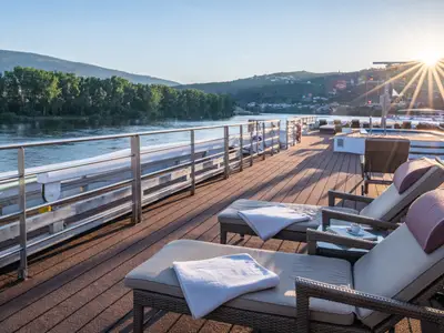 Blick über das Sonnendeck des Flusskreuzfahrtschiffs Douro Queen. Komfortable Liegestühle stehen bereit, während die untergehende Sonne den Douro-Fluss und die umliegenden grünen Hügel beleuchtet. Lange Schatten ziehen sich über das Deck.