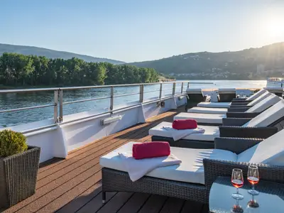Das Sonnendeck der Douro Queen mit Liegestühlen, roten Handtüchern und Weingläsern. Blick auf den Fluss Douro und grüne Hügel unter strahlender Sonne.