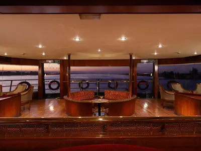 Ansicht der eleganten Lounge der Jaz Nile Monarch. Man sieht bequeme Sitzgruppen aus Holz und Polstern sowie große Fenster mit Blick auf einen Fluss bei Sonnenuntergang. Deckenleuchten erhellen den Raum.