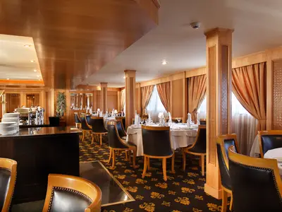 Innenansicht des Restaurants der Jaz Nile Monarch. Festlich gedeckte Tische, bequeme Stühle, Holzwände, gemusterter Teppich und eine Buffetstation sind zu sehen.