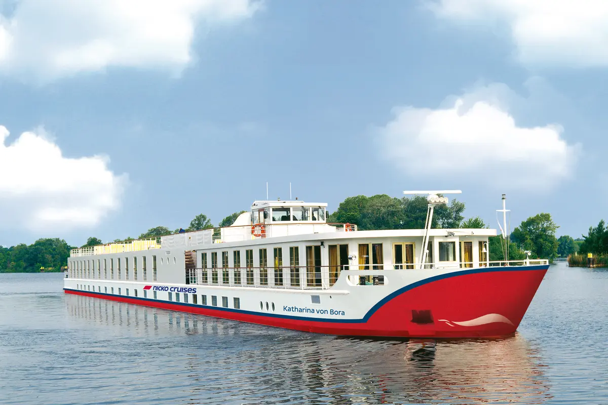 Die MS Katharina von Bora, ein weiß-rotes Flussschiff mit blauen Akzenten, fährt auf einem Fluss an bewachsenen Ufern vorbei. Der Himmel ist leicht bewölkt.
