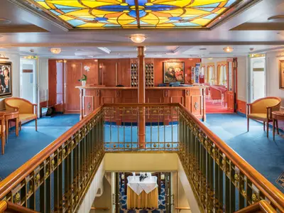 Foyer des Flusskreuzfahrtschiffs Katharina von Bora. Zu sehen sind der Holztresen der Rezeption, blaue Teppichböden, eine kunstvolle Buntglasdecke und ein Treppenhaus mit elegantem Geländer.