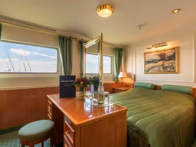 Helle Kabine auf dem Flusskreuzfahrtschiff Katharina von Bora. Doppelbett, Schreibtisch mit Blumen und Ausblick durch die Fenster auf Schiffsmasten im Hafen.