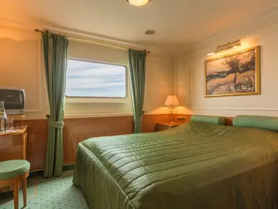 Eine helle Außenkabine mit einem großen, grünen Doppelbett, einem Fenster mit Meerblick, einem Fernseher und einem Kunstgemälde an der Wand. Neben dem Fenster befinden sich grüne Vorhänge, und ein kleiner Tisch mit einer Wasserflasche und Gläsern steht links.