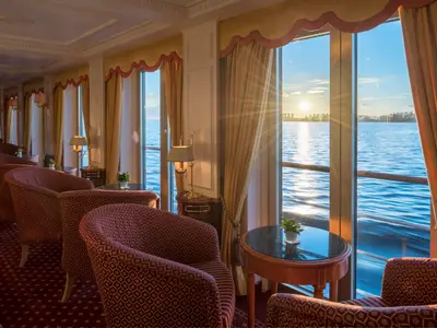 Lounge der Katharina von Bora. Rote Sessel und Tische stehen an großen Fenstern. Blick auf einen Fluss bei Sonnenuntergang, dessen Licht auf dem Wasser glitzert.