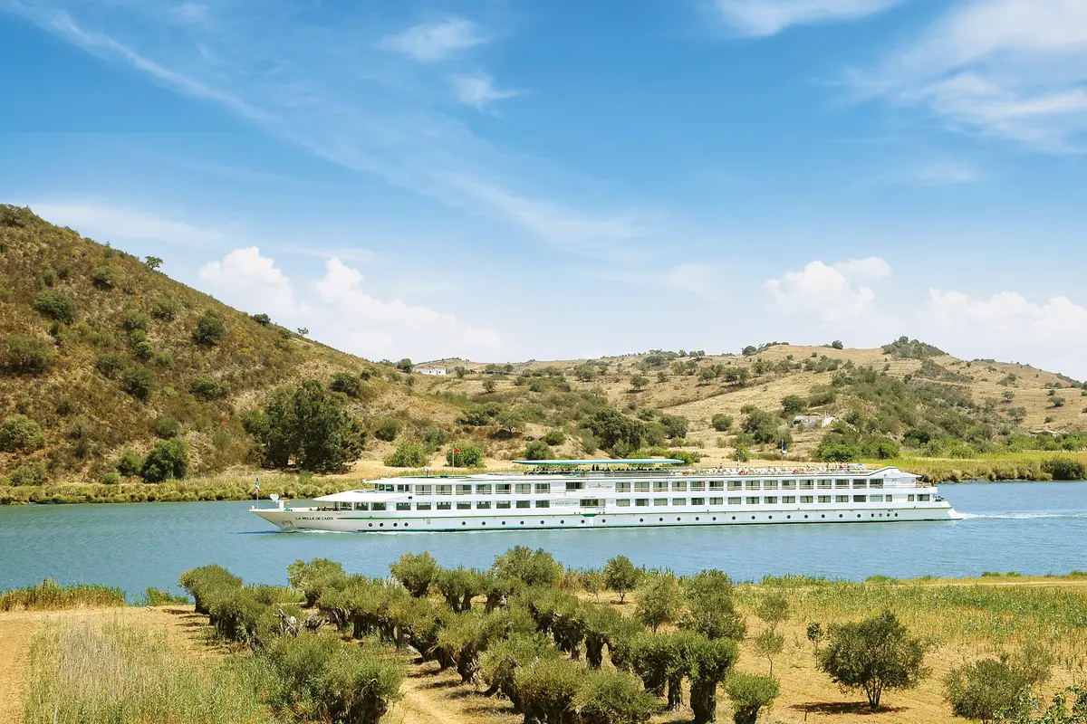 Flussschiff La Belle de Cadix von CroisiEurope fährt auf einem Fluss. Im Vordergrund Olivenbäume, im Hintergrund trockene, hügelige Landschaft und blauer Himmel.