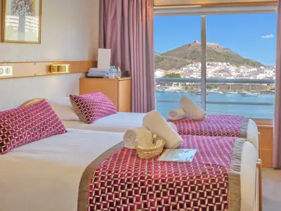 Helle Kreuzfahrt-Außenkabine mit zwei Einzelbetten und pinken Tagesdecken. Großes Fenster mit Blick auf einen Hafen mit Booten, weiße Häuser und einen bergigen Hintergrund mit einer Festung. Auf dem Bett liegen Pflegeprodukte und gerollte Handtücher bereit.