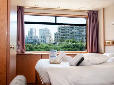 Kabine des Flusskreuzfahrtschiffs La Belle de Cadix. Bett mit Handtüchern und Fernbedienung. Große Fensterfront mit Blick auf Stadtsilhouette und Flusslandschaft.