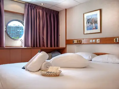 Eine Kabine an Bord der La Belle de Cadix: Weißes Bett mit Handtüchern, Bullauge mit Blick auf Gebäude. Darüber ein maritimes Gemälde und Leselampen.