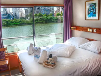 Eine helle Kabine an Bord der La Belle de Cadix zeigt ein gemütliches Bett mit weißen Laken und Handtüchern. Durch das große Fenster ist ein Fluss und die städtische Umgebung zu sehen.