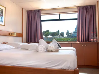 Helle Außenkabine mit zwei Einzelbetten und Fensterblick auf grüne Landschaft. Frische Handtücher und Wasserflaschen auf der Fensterbank. Ein Bild an der Wand.