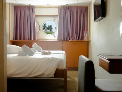 Blick in eine Kabine der La Belle de Cadix: Zwei Betten mit weißen Handtüchern, ein Bullauge mit Aussicht auf Bäume. Rechts ein Fernseher und ein Schreibtisch sind zu sehen.