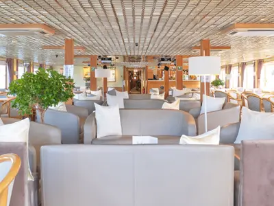 Blick in die helle Lounge der La Belle de Cadix mit komfortablen, hellgrauen Sofas und Sesseln sowie weißen Kissen. Im Hintergrund sind Fenster und eine Bar erkennbar.