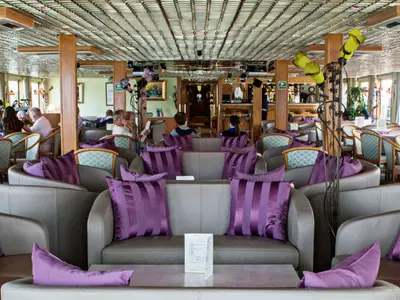 Einrichtung der Lounge auf der La Belle de Cadix, mit hellgrauen Sesseln, lila Kissen und entspannten Gästen. Eine Bar ist im Hintergrund sichtbar.
