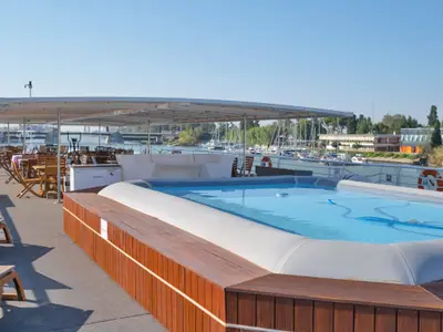 Sonnendeck der La Belle de Cadix mit Pool, Liegen und überdachtem Essbereich. Im Hintergrund Yachthafen, Gebäude und eine Brücke am Fluss.