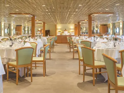 Heller Speisesaal der La Belle de Cadix mit vielen gedeckten Tischen und hellgrünen Stühlen. Bullaugen und ein Buffet mit Obst sind im Hintergrund erkennbar.
