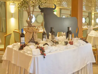 Im Speisesaal der La Belle de Cadix steht ein festlicher Tisch mit Weinflaschen, Trauben und einem schwarzen Stier. Weitere Tische und Bullaugen sind im Hintergrund sichtbar.
