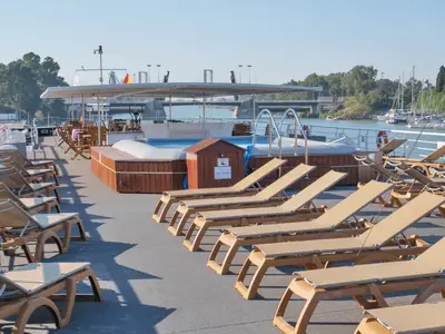 Sonnendeck der La Belle de Cadix mit Reihen beiger Sonnenliegen und einem Pool. Im Hintergrund sind ein Fluss, eine Brücke und grüne Ufer mit weiteren Booten erkennbar.