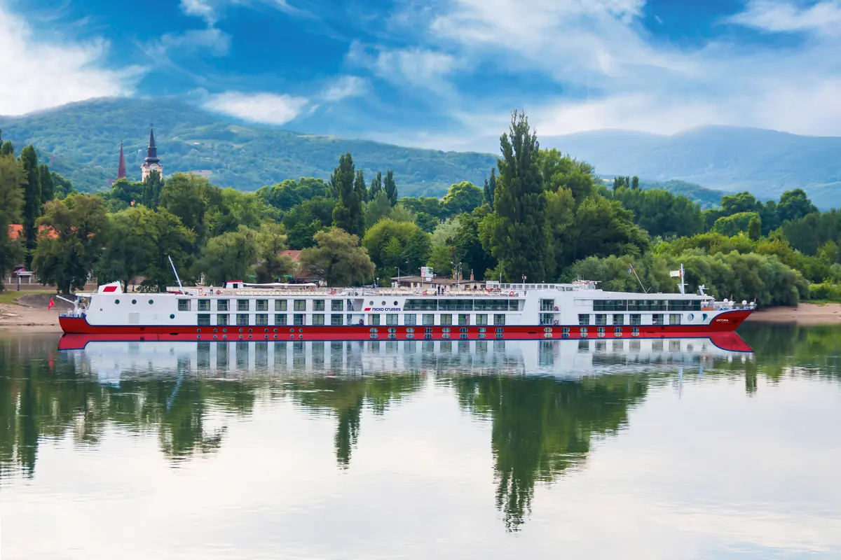 Das Flusskreuzfahrtschiff Maxima von nicko cruises auf einem Fluss, umgeben von grünen Ufern und Hügeln. Blauer Himmel. Schiffsspiegelung im Wasser.