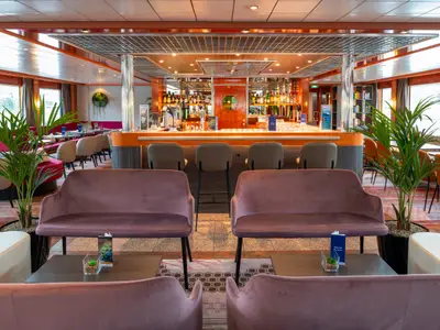 Der Bar- und Lounge-Bereich des Flusskreuzfahrtschiffs Maxima zeigt eine orange Bar, Sitzgruppen und Pflanzen. Große Fenster geben den Blick frei.