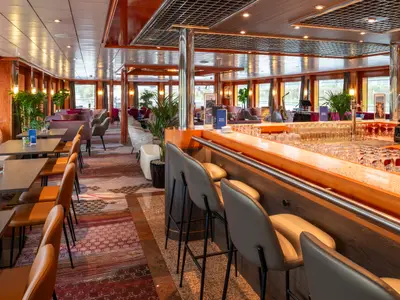 Helle Bar und Lounge auf der Maxima. Rechts Theke mit Hockern, Gläsern. Im Hintergrund Sitzgruppen, Fenster, ein Saxophon. Das moderne Interieur der Maxima ist einladend.