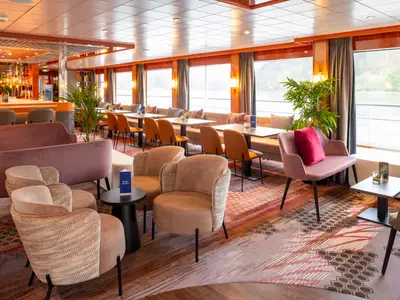 Modern eingerichtete Lounge auf der Maxima. Eine Bar, gemütliche Sitzgruppen und Tische an Panoramafenstern mit Flussblick.