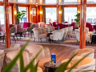 Die stilvolle Lounge des nicko cruises Flusskreuzfahrtschiffs Maxima. Helle und farbige Sessel an Tischen, umgeben von großen Fenstern. Einladender Bereich an Bord der Maxima.