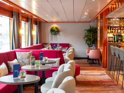 Die Lounge des Flusskreuzfahrtschiffs Maxima. Fuchsiafarbene Sitzbänke an Fenstern mit Tischen. Rechts eine Bar mit Hockern. Moderne und freundliche Inneneinrichtung.