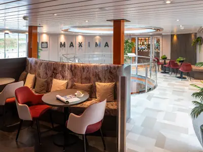 Lounge auf dem Flusskreuzfahrtschiff Maxima: Moderne Sitzgelegenheiten, Tische, Fenster. Im Hintergrund Wendeltreppe, Bar und 'MAXIMA' Schriftzug.