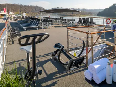 Das Oberdeck der Maxima mit Fitnessgeräten, Handtüchern und Wasserflaschen. Im Hintergrund sind Liegestühle, Tische, ein Sonnendach, ein Fluss und bewaldete Hügel zu sehen.