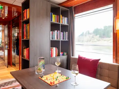 Loungebereich der nicko cruises Maxima. Tisch mit Brettspiel, zwei Weingläsern. Bücherregal. Fenster mit Flussblick. Sofa mit rotem Kissen.