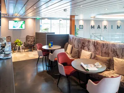 Lounge und Buffetbereich des Flusskreuzfahrtschiffs Maxima. Moderne Sitzecken mit Tischen und Stühlen. An der Wand steht der Name Maxima.