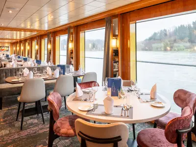 Das Restaurant auf dem Flusskreuzfahrtschiff Maxima. Die Tische sind gedeckt, große Fenster bieten Ausblick auf den Fluss und das bewaldete Ufer.