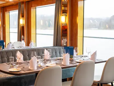 Restaurant auf der maxima. Gedeckte Tische mit Geschirr, Gläsern und Besteck, gepolsterte Sitzbänke und Stühle sind sichtbar. Große Fenster zeigen die Flusslandschaft draußen.