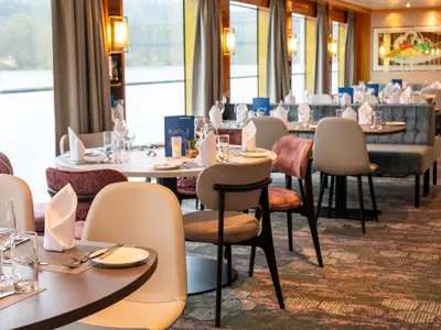 Restaurant der Maxima. Tische mit Besteck und Gläsern, Stühle und Sitzbänke. Fenster mit Flussblick. Warme Lichter, Wandkunst.