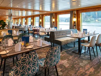 Das helle Restaurant des Flusskreuzfahrtschiffs Maxima zeigt eingedeckte Tische, moderne Stühle mit Muster und bequeme Sitzbänke. Große Fenster erlauben den Blick auf das Ufer.