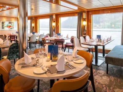 Das Restaurant der Maxima mit gedeckten Tischen und Panoramafenstern bietet einen weiten Blick auf die Flusslandschaft. Eine moderne Atmosphäre prägt das Innere der Maxima.
