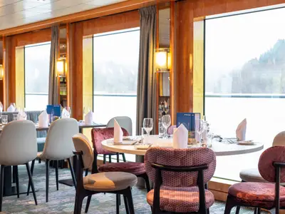 Helles Restaurant an Bord der Maxima, mit gedeckten Tischen und modernen Stühlen. Große Fenster zeigen eine bewaldete Flusslandschaft. Das Interieur der Maxima ist mit Holz und Lichtakzenten gestaltet.
