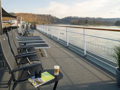 Sonnendeck der Maxima. Liegestühle, Tisch mit Fernglas, Buch und Latte Macchiato. Blick auf Fluss und bewaldete Ufer. Ein ruhiger Tag an Bord der Maxima.