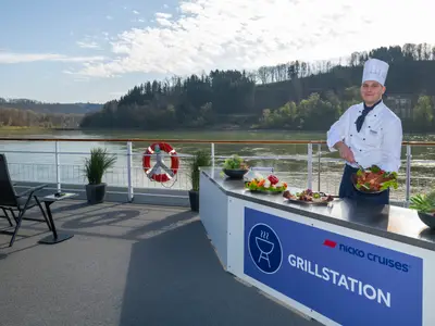 Koch an der Grillstation auf der Maxima. Serviert Grillgut. Fluss und Wald im Hintergrund. Rettungsboje, Deckstühle sichtbar.