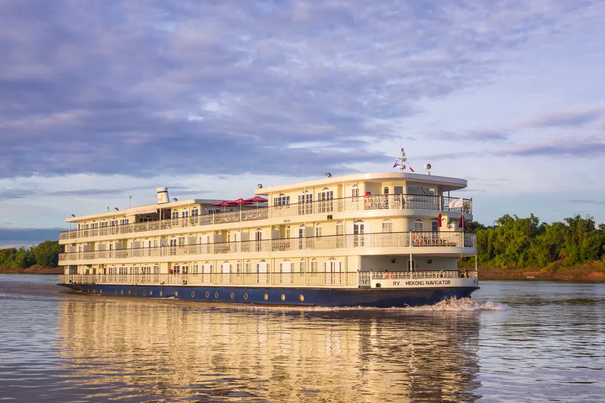 Das weiße Flussschiff RV Mekong Navigator mit mehreren Decks und goldenen Geländern fährt auf einem breiten Fluss. Grüne Ufervegetation ist im Hintergrund sichtbar, der Himmel ist bewölkt, und die Sonne reflektiert golden auf dem Wasser.