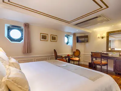Eine elegant eingerichtete Kabine des Flusskreuzfahrtschiffs Mekong Navigator. Sie zeigt ein großes Bett, Bullaugen, eine Sitzecke und einen Schminktisch mit Spiegel.