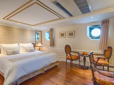 Geräumige Kabine des Mekong Navigator mit einem großen Bett, einer eleganten Sitzgruppe, Bullauge und Holzboden. Die Decke ist goldverziert.