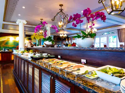 Ein luxuriöses Buffet mit frischen Salaten, Aufschnitt und Dips. Elegante Dekoration mit lila Orchideen und Kronleuchtern. Ein Crew-Mitglied ist im Hintergrund zu sehen.