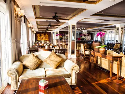 Lounge des Flusskreuzfahrtschiffs Mekong Navigator mit Sofas, Holzfußboden, Bar und goldenem Wandrelief. Helles Tageslicht durch große Fenster.