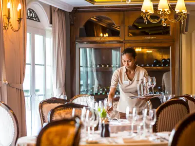 Im Restaurant des Flusskreuzfahrtschiffs Mekong Navigator deckt eine Mitarbeiterin Tische. Sie hält ein Tablett mit Gläsern. Die elegante Einrichtung umfasst Holzmöbel und große Fenster.