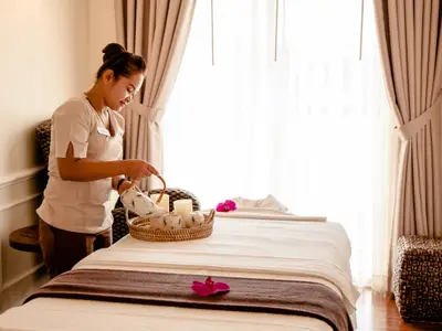 Spa-Mitarbeiterin gießt Tee in Tasse auf Tablett. Kerzen und pinke Orchideenblüten dekorieren das Massagebett in einem hellen, entspannten Spa-Raum.