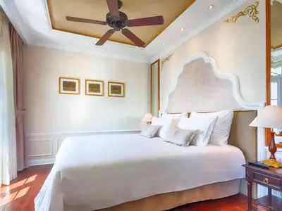 Helle, elegante Suite mit großem, weiß bezogenem Bett, kunstvollem Kopfteil, Deckenventilator und Vorhängen am Fenster.
